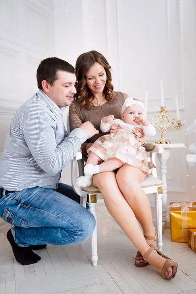 Baby girl wearing cute dress and headband with mother and father, near christmas tree in festively decorated room with garland of lights. Warm beige and gold colors of christmas and new year atmospher