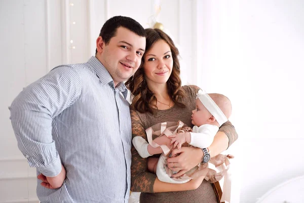 Baby girl wearing cute dress and headband with mother and father, near christmas tree in festively decorated room with garland of lights. Warm beige and gold colors of christmas and new year atmospher