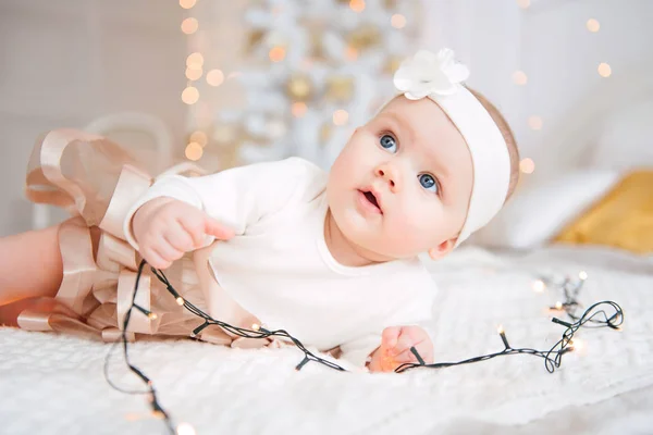 Bébé fille portant une robe mignonne et un bandeau, se trouve sur une couverture blanche dans une chambre décorée avec une guirlande de lumières. Avec des montres surprises dans la caméra, sur un fond un ensemble de feux lumineux. Que ce soit chaud — Photo