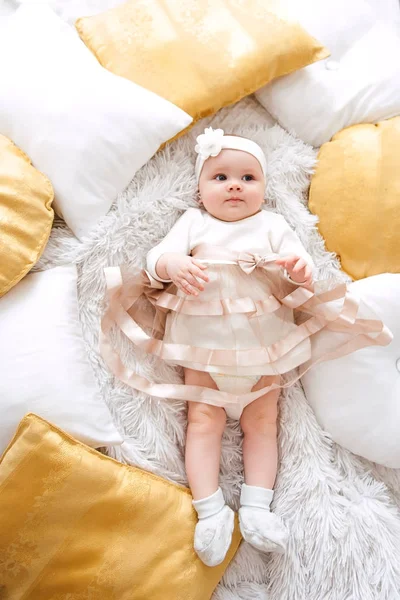 Baby girl wearing cute dress and headband, lies on a white cover in festively decorated room with garland of lights. With surprise watches in the camera, on a background a set of bright fires. Warm be