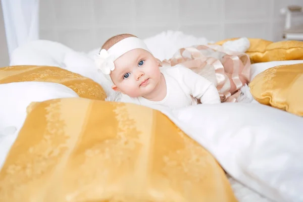 Baby girl wearing cute dress and headband, lies on a white cover in festively decorated room with garland of lights. With surprise watches in the camera, on a background a set of bright fires. Warm be