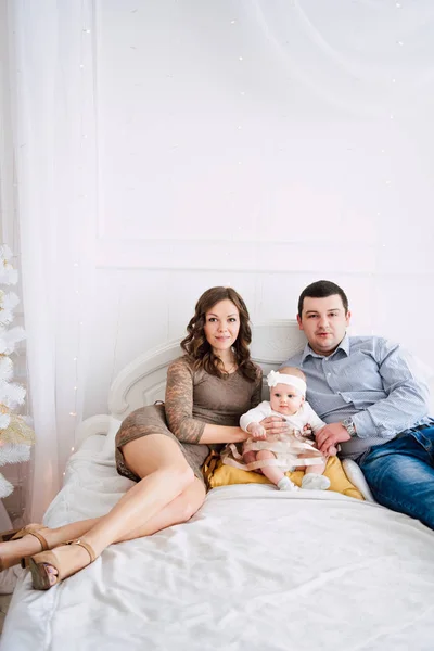 Baby girl wearing cute dress and headband with mother and father, near christmas tree in festively decorated room with garland of lights. Warm beige and gold colors of christmas and new year atmospher