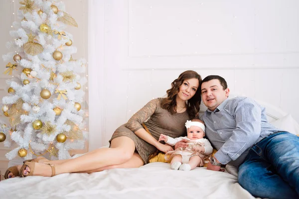 Niña vistiendo lindo vestido y diadema con la madre y el padre, cerca del árbol de Navidad en la habitación festivamente decorada con guirnalda de luces. Los colores cálidos de color beige y oro de la Navidad y el ambiente de año nuevo — Foto de Stock