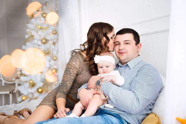 Baby girl wearing cute dress and headband with mother and father, near christmas tree in festively decorated room with garland of lights. Warm beige and gold colors of christmas and new year atmospher