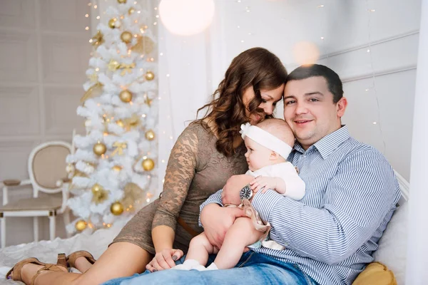 Mädchen in süßem Kleid und Stirnband mit Mutter und Vater, in der Nähe des Weihnachtsbaums in festlich geschmücktem Raum mit Lichterkranz. warme Beige- und Goldtöne der Weihnachts- und Neujahrsatmosphäre — Stockfoto