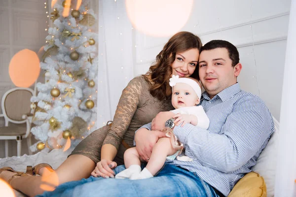 Baby girl wearing cute dress and headband with mother and father, near christmas tree in festively decorated room with garland of lights. Warm beige and gold colors of christmas and new year atmospher