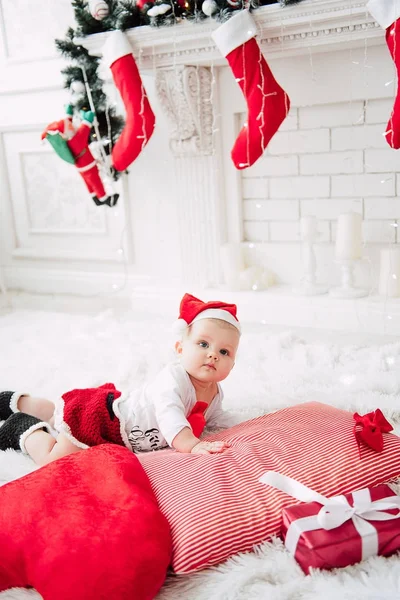 Baby girl wearing cute red dress and santa hat, near christmas tree in festively decorated room with garland of lights. White and red colors of christmas and new year atmosphere. Concept of a happy fa