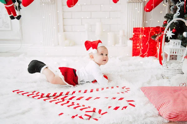お祝いを着てかわいい赤いドレス、サンタ帽子、クリスマス ツリーの近くの女の赤ちゃんは、ライトのガーランドと部屋を飾った。クリスマスとお正月の雰囲気の白と赤の色。幸せそうな顔の概念 — ストック写真