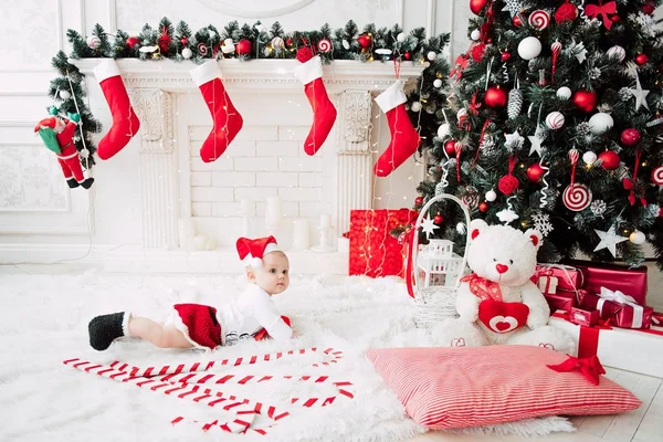 Niña vistiendo lindo vestido rojo y sombrero de santa, cerca del árbol de Navidad en la habitación festivamente decorada con guirnalda de luces. Los colores blancos y rojos de la Navidad y la atmósfera de año nuevo. Concepto de un fa feliz — Foto de Stock