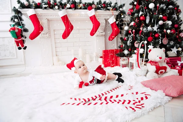 Baby girl wearing cute red dress and santa hat, near christmas tree in festively decorated room with garland of lights. White and red colors of christmas and new year atmosphere. Concept of a happy fa