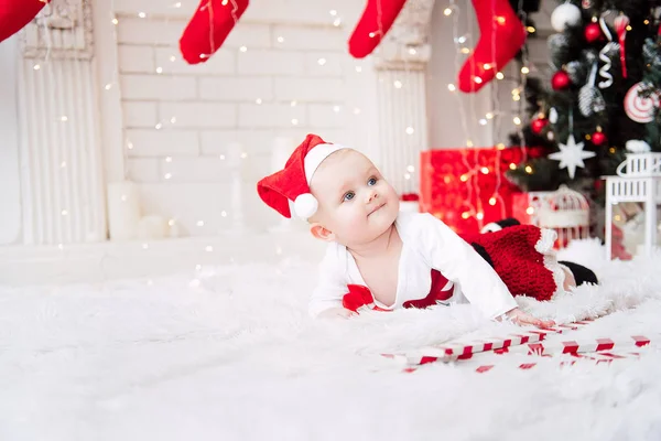 Baby flicka klädd i söt röd klänning och santa hatt, nära julgran i festligt inrett rum med krans av lampor. Vita och röda färger av jul och nyår atmosfär. Begreppet en glad fa — Stockfoto