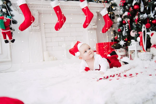 Baby flicka klädd i söt röd klänning och santa hatt, nära julgran i festligt inrett rum med krans av lampor. Vita och röda färger av jul och nyår atmosfär. Begreppet en glad fa — Stockfoto