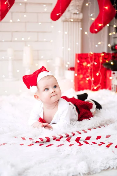 Baby flicka klädd i söt röd klänning och santa hatt, nära julgran i festligt inrett rum med krans av lampor. Vita och röda färger av jul och nyår atmosfär. Begreppet en glad fa — Stockfoto