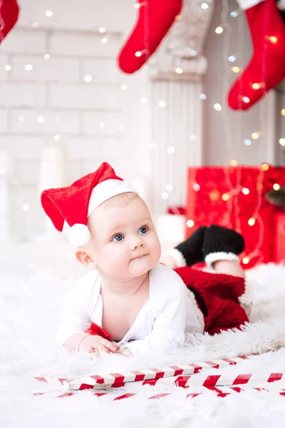 Niña vistiendo lindo vestido rojo y sombrero de santa, cerca del árbol de Navidad en la habitación festivamente decorada con guirnalda de luces. Los colores blancos y rojos de la Navidad y la atmósfera de año nuevo. Concepto de un fa feliz — Foto de Stock