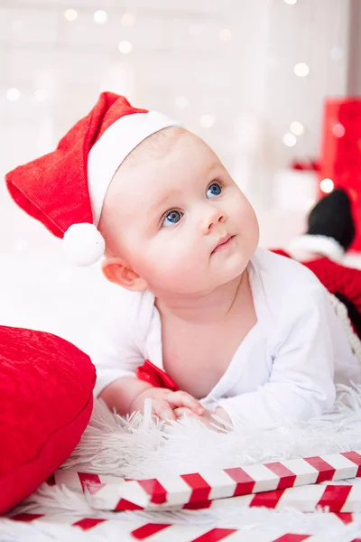 Niña vistiendo lindo vestido rojo y sombrero de santa, cerca del árbol de Navidad en la habitación festivamente decorada con guirnalda de luces. Los colores blancos y rojos de la Navidad y la atmósfera de año nuevo. Concepto de un fa feliz — Foto de Stock