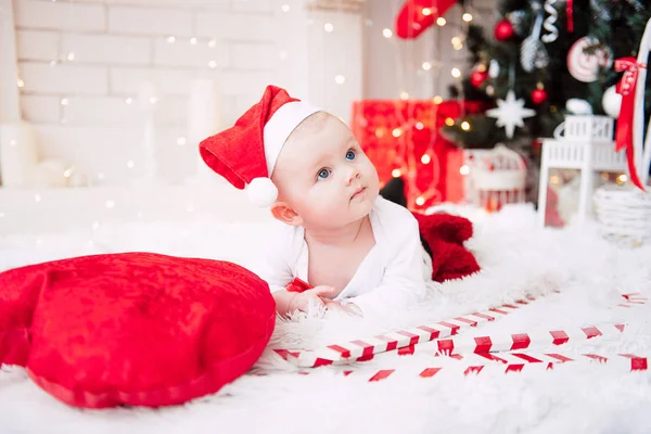 Baby flicka klädd i söt röd klänning och santa hatt, nära julgran i festligt inrett rum med krans av lampor. Vita och röda färger av jul och nyår atmosfär. Begreppet en glad fa — Stockfoto