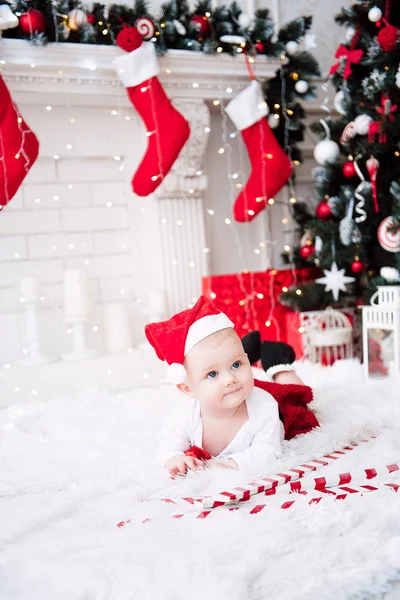 Niña vistiendo lindo vestido rojo y sombrero de santa, cerca del árbol de Navidad en la habitación festivamente decorada con guirnalda de luces. Los colores blancos y rojos de la Navidad y la atmósfera de año nuevo. Concepto de un fa feliz — Foto de Stock