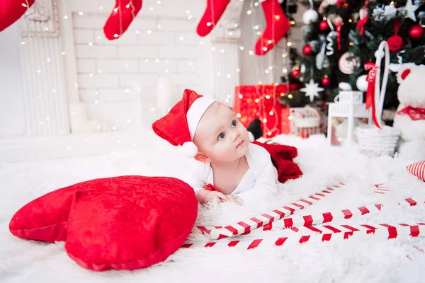 Baby girl wearing cute red dress and santa hat, near christmas tree in festively decorated room with garland of lights. White and red colors of christmas and new year atmosphere. Concept of a happy fa