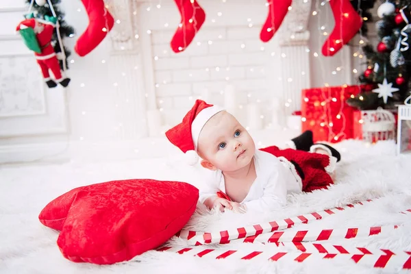 Baby girl wearing cute red dress and santa hat, near christmas tree in festively decorated room with garland of lights. White and red colors of christmas and new year atmosphere. Concept of a happy fa