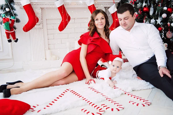 Baby flicka klädd i söt röd klänning och santa hatt med mor i röd klänning och far, nära julgran i festligt inrett rum med krans av lampor. Vita och röda färger av jul och nya ni — Stockfoto
