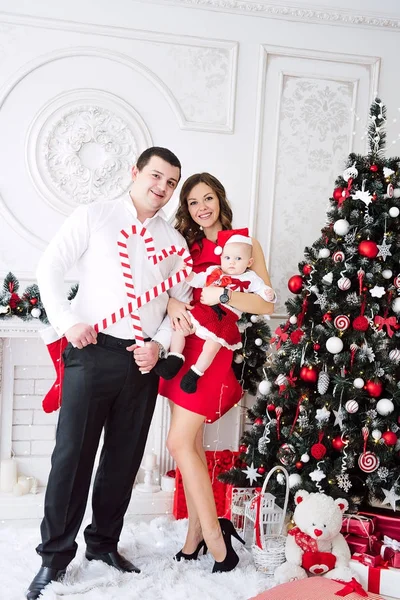 Baby girl wearing cute red dress and santa hat with mother in red dress and father, near christmas tree in festively decorated room with garland of lights. White and red colors of christmas and new ye