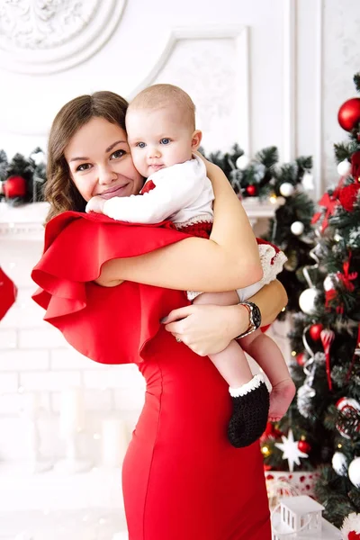 Baby girl wearing cute red dress and santa hat with mother in red dress, near christmas tree in festively decorated room with garland of lights. White and red colors of christmas and new year atmosphe