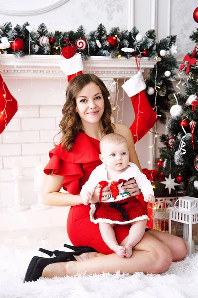 Baby flicka klädd i söt röd klänning och santa hatt med mor i röd klänning, nära julgran i festligt inrett rum med krans av lampor. Vita och röda färger av jul och nyår atmo — Stockfoto