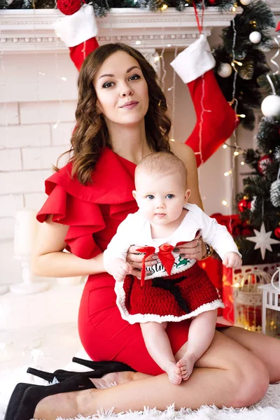 Baby girl wearing cute red dress and santa hat with mother in red dress, near christmas tree in festively decorated room with garland of lights. White and red colors of christmas and new year atmosphe