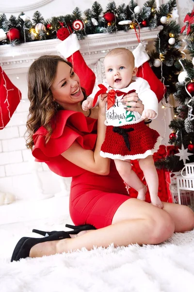 Baby girl wearing cute red dress and santa hat with mother in red dress, near christmas tree in festively decorated room with garland of lights. White and red colors of christmas and new year atmosphe