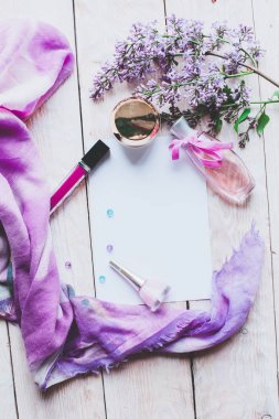 Düz yatıyordu, üst görünüm yaylı masa çerçevesi. Ahşap zemin üzerinde kozmetik, ruj ve leylak çiçekleri olan kadınsı masa çalışma alanı.