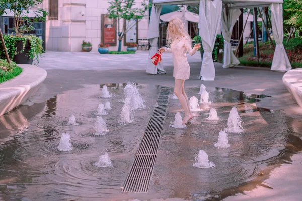 Beautiful young blonde smiling happy woman with red lips in red shoes in a white dress play near a fountain in a summer day — Stock Photo, Image