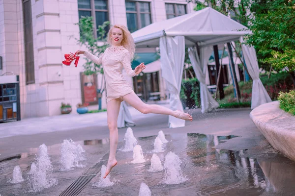 Bella giovane bionda sorridente donna felice con le labbra rosse in scarpe rosse in un vestito bianco giocare vicino a una fontana in una giornata estiva — Foto Stock