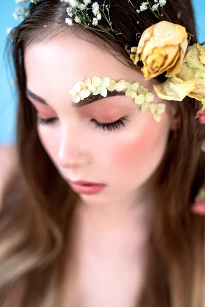 Cosméticos e manicure. Retrato close-up de mulher atraente com flores secas em seu rosto e cabelo, cor pastel, maquiagem perfeita e pele no fundo azul. Retrato retocado de primavera fresco, na moda — Fotografia de Stock