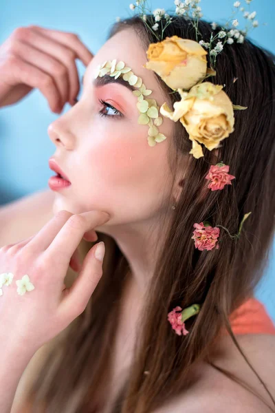 Cosméticos e manicure. Retrato close-up de mulher atraente com flores secas em seu rosto e cabelo, cor pastel, maquiagem perfeita e pele no fundo azul. Retrato retocado de primavera fresco, na moda — Fotografia de Stock