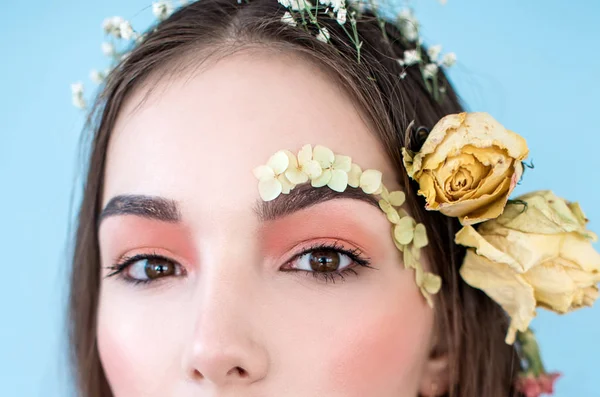 Cosméticos y manicura. Retrato de cerca de una mujer atractiva con flores secas en la cara y el cabello, color pastel, maquillaje perfecto y piel sobre fondo azul. Retrato retocado de primavera fresco, moderno — Foto de Stock