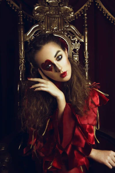 Woman dressed as the queen of hearts, creative make up with big heart on eye, girl sitting in the throne, hi-end retouched portrait. Conceptual photo