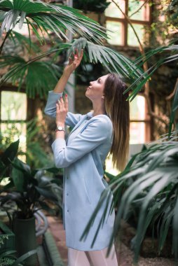 Genç, zarif, pozitif kız botanik bahçesinde mavi ceketli poz veriyor..