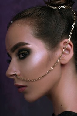 Havalı makyajlı bir kız stüdyoda poz veriyor, mat dudaklar ve tırnaklar, rengârenk gözler, dumanlı altın gözler, güzel bir makyaj ve mor arkaplanda parlak mor bir arka planla burundan piercing zinciri. Tatil disko kutlaması