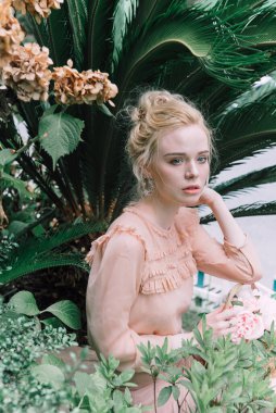 Şehvetli genç sarışın güzel ve zarif bir kadın kayaya oturmuş Çin 'deki tropik yeşil orman palmiyesinden kameraya bakıyor..