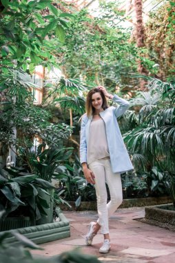 Genç, zarif, pozitif kız botanik bahçesinde mavi ceketli poz veriyor..