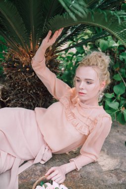 Şehvetli genç sarışın güzel ve zarif bir kadın kayaya oturmuş Çin 'deki tropik yeşil orman palmiyesinden kameraya bakıyor..