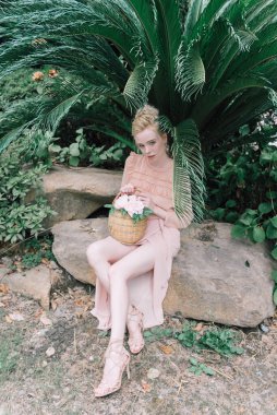 Şehvetli genç sarışın güzel ve zarif bir kadın kayaya oturmuş Çin 'deki tropik yeşil orman palmiyesinden kameraya bakıyor..