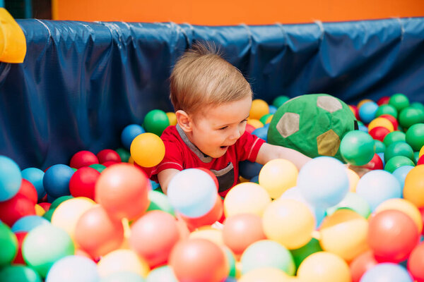 Happy laughing boy 1-2 years old having fun in ball pit on birthday party in kids amusement park and indoor play center. Child playing with colorful balls in playground ball pool. Activity toys for little kid.