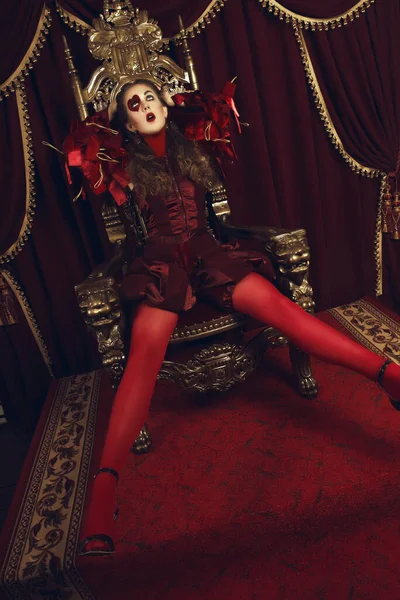 Woman dressed as the queen of hearts, creative make up with big heart on eye, girl sitting in the throne, hi-end retouched portrait. Conceptual photo
