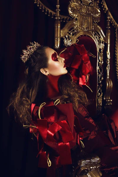 Woman dressed as the queen of hearts, creative make up with big heart on eye, girl sitting in the throne, hi-end retouched portrait. Conceptual photo