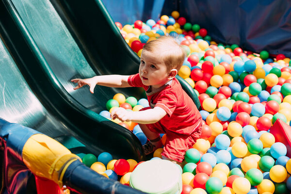 Happy laughing boy 1-2 years old having fun in ball pit on birthday party in kids amusement park and indoor play center. Child playing with colorful balls in playground ball pool. Activity toys for little kid.