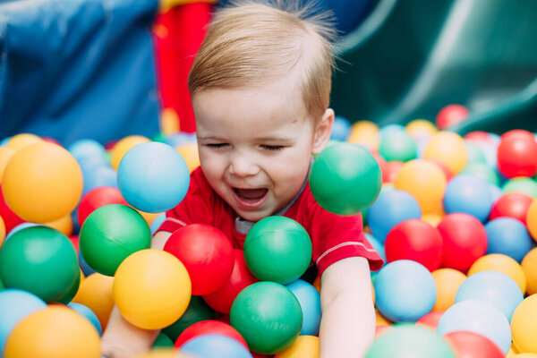 Happy laughing boy 1-2 years old having fun in ball pit on birthday party in kids amusement park and indoor play center. Child playing with colorful balls in playground ball pool. Activity toys for little kid.