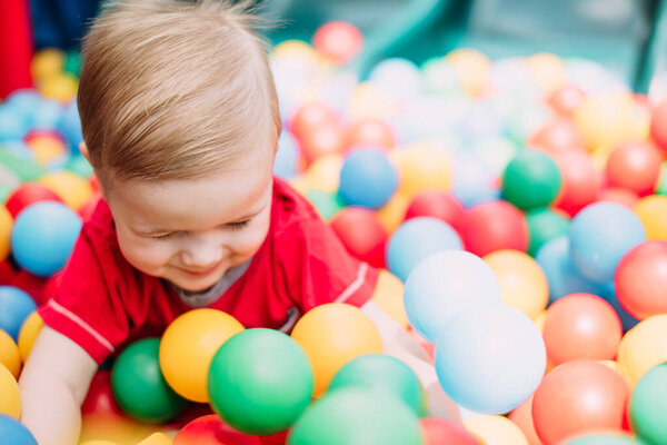 Happy laughing boy 1-2 years old having fun in ball pit on birthday party in kids amusement park and indoor play center. Child playing with colorful balls in playground ball pool. Activity toys for little kid.