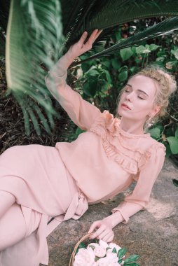 Şehvetli genç sarışın güzel ve zarif bir kadın kayaya oturmuş Çin 'deki tropik yeşil orman palmiyesinden kameraya bakıyor..