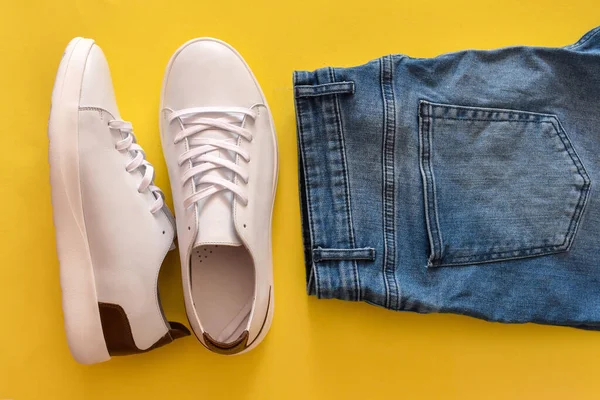 Set of white sneakers and blue jeans on yellow background.